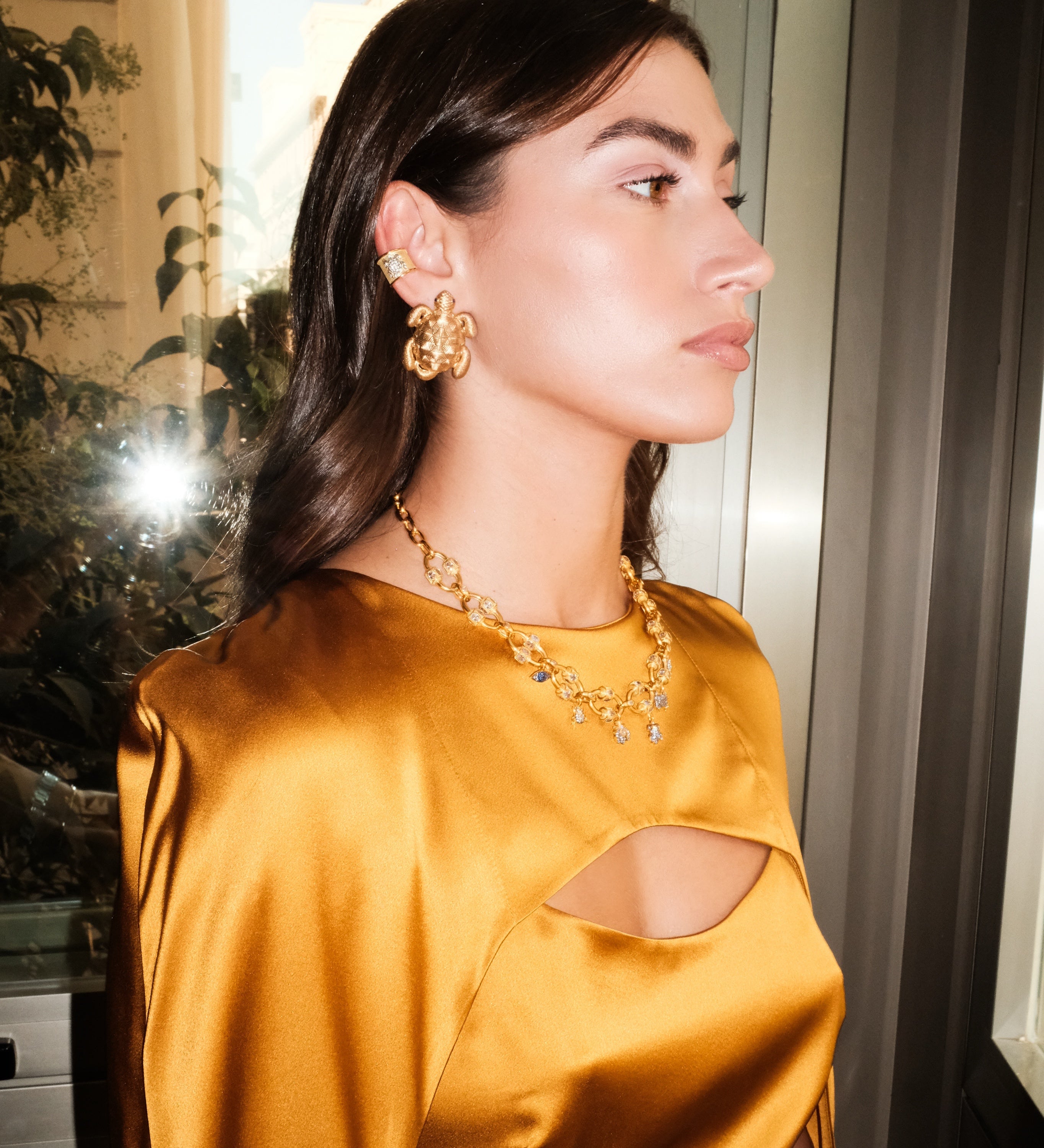 Woman wearing a mustard yellow satin dress with cut-out design, gold earrings, and necklace.