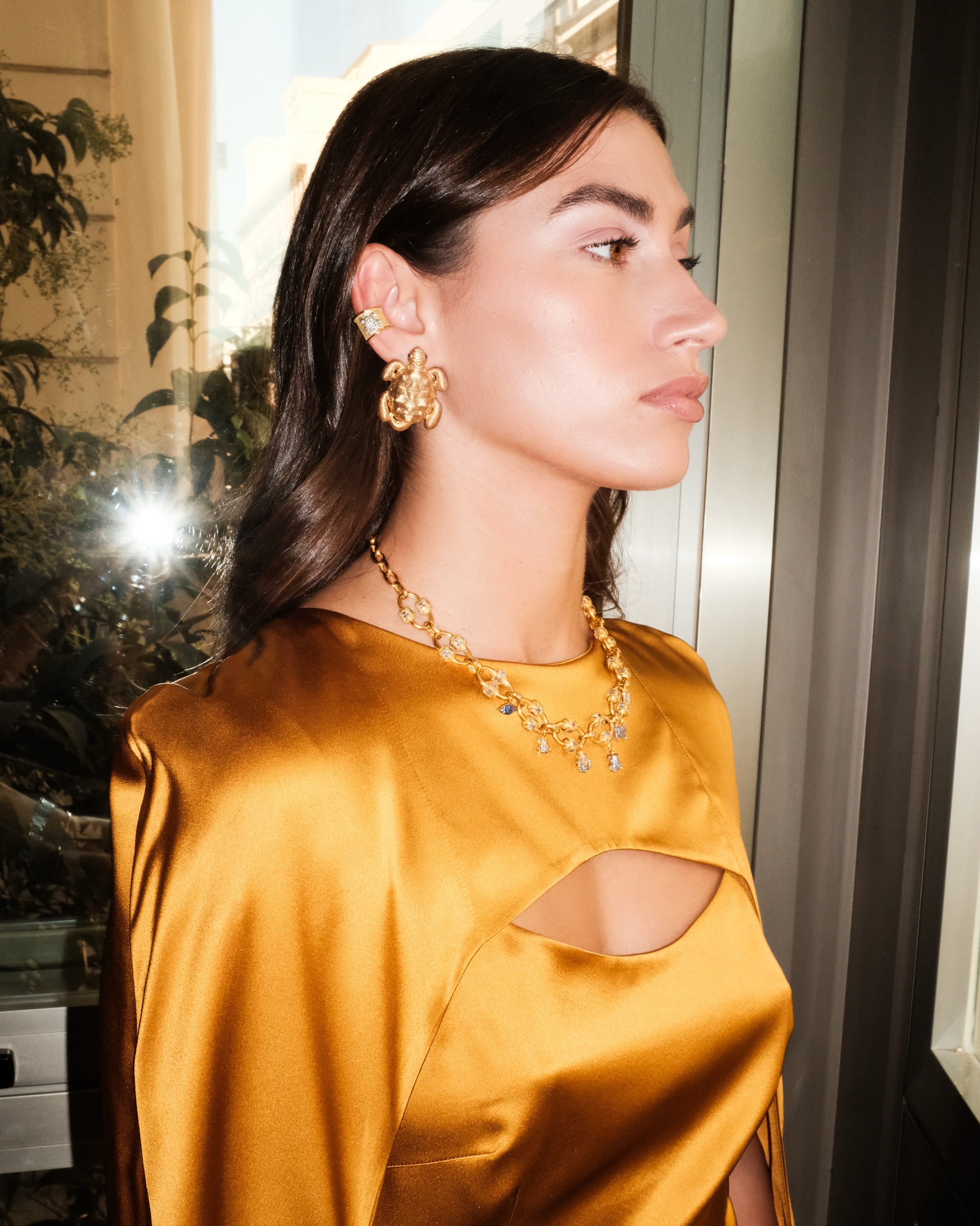 Woman wearing a mustard yellow satin dress with cut-out design, gold earrings, and necklace.